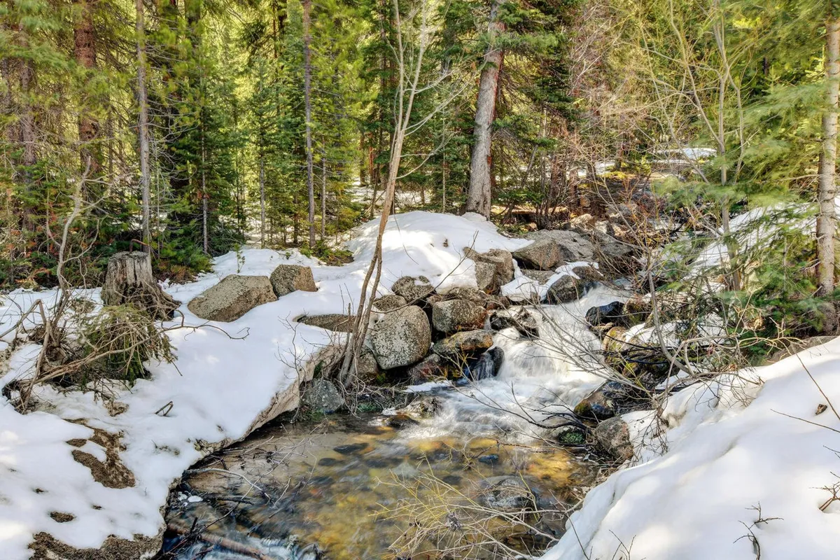 A tranquil, snow-covered mountain landscape featuring a gently flowing stream, with tall evergreens surrounding the scene and rocks blanketed in fresh snow. The environment evokes a connection to the outdoors and the Winter Park mountain setting.