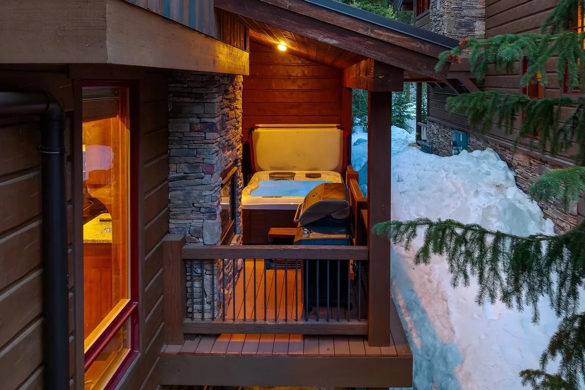 An inviting outdoor terrace featuring a bubbling hot tub, a gas grill, and outdoor seating on a wooden deck bordered by a stone wall. Snow blankets the surroundings, hinting at year-round enjoyment, while the hot tub steams beneath a starlit sky.