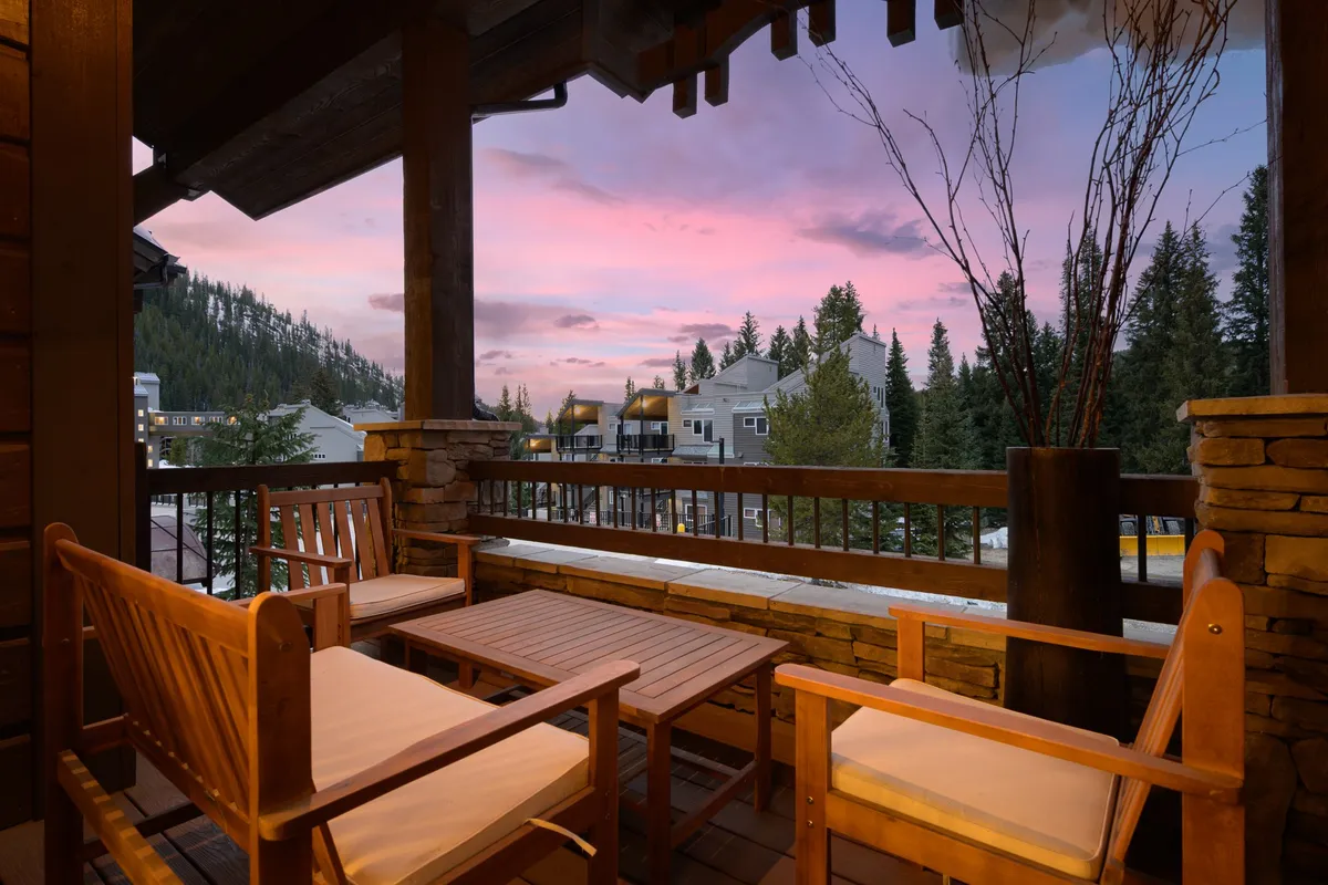 A tranquil outdoor patio with wooden furniture overlooking a breathtaking mountain view, set under a vibrant sunset sky. The scene invites relaxation and socializing amid natural beauty.