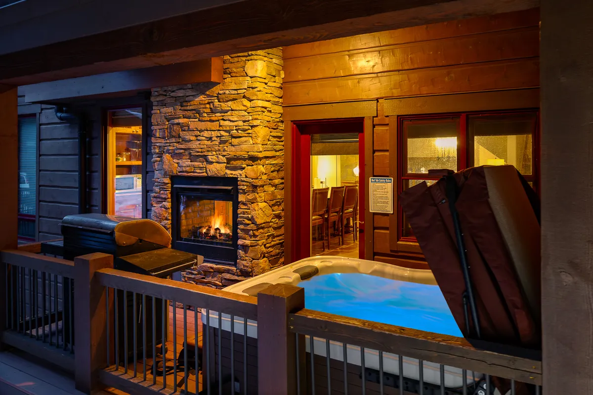 A cozy indoor space showcasing a hot tub set against wooden walls, a glowing fireplace, and an adjacent dining area. Large windows reveal falling snow, further blending the indoor and outdoor experience.