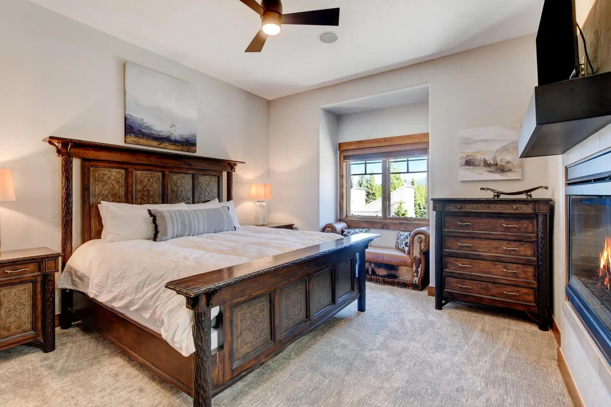 A warmly lit bedroom with a wooden bedframe, window offering a view, a fireplace opposite the bed, and a wooden dresser. The room exudes a tranquil mountain ambiance, with plush bedding and cozy finishes.