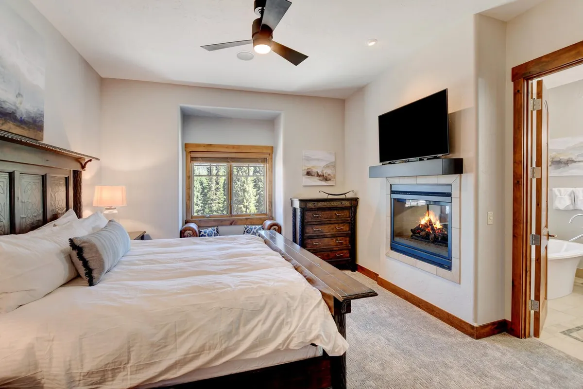 A serenely decorated bedroom with a bed, fireplace, large window, dresser, and HDTV. Sunlight pours in, creating a calm, relaxing setting ideal for unwinding and private entertainment.