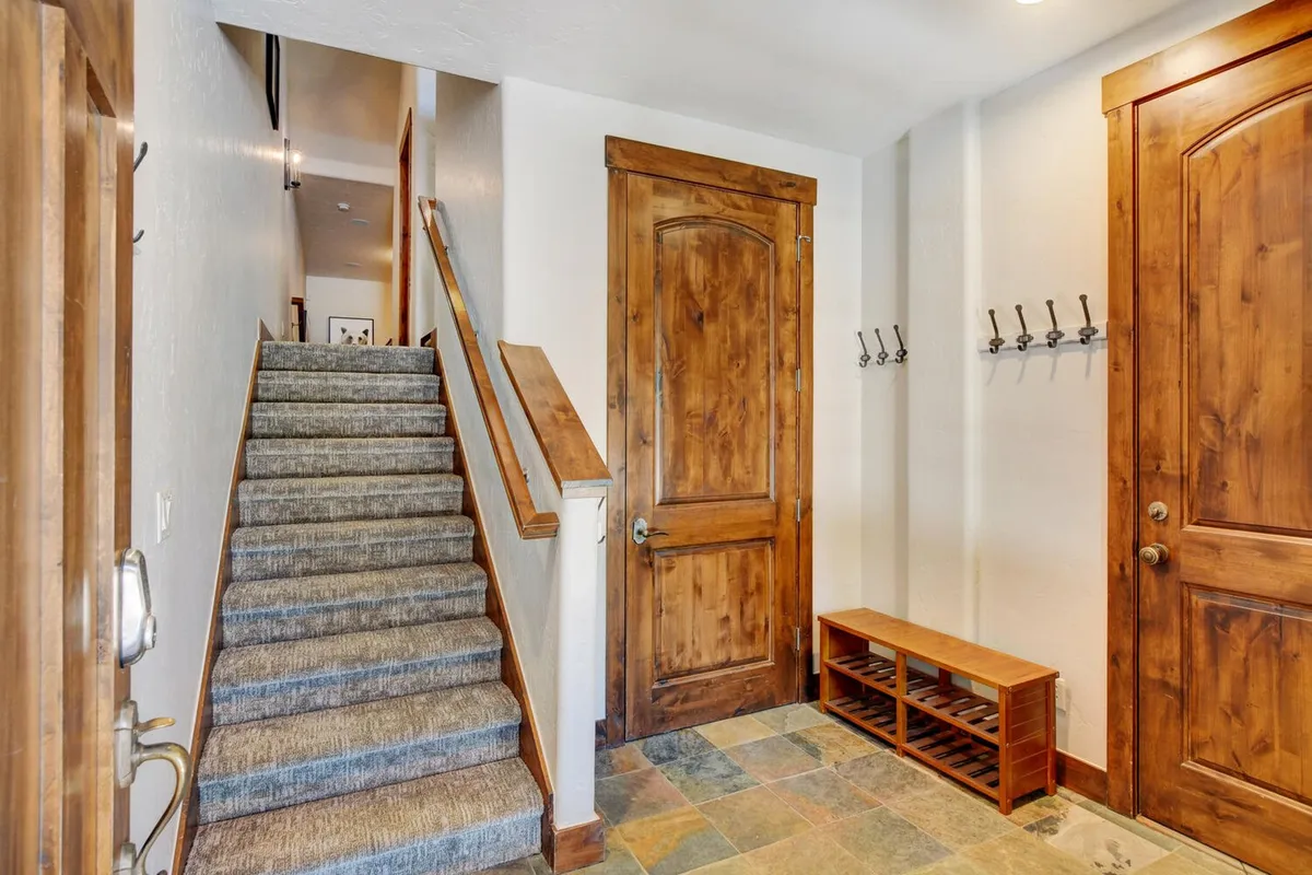 A cozy entry space featuring a staircase, wooden door, entry bench, and coat hooks, designed for welcoming guests and storing outdoor gear after a day of activities. The warm tones and practical layout emphasize both hospitality and convenience for families.