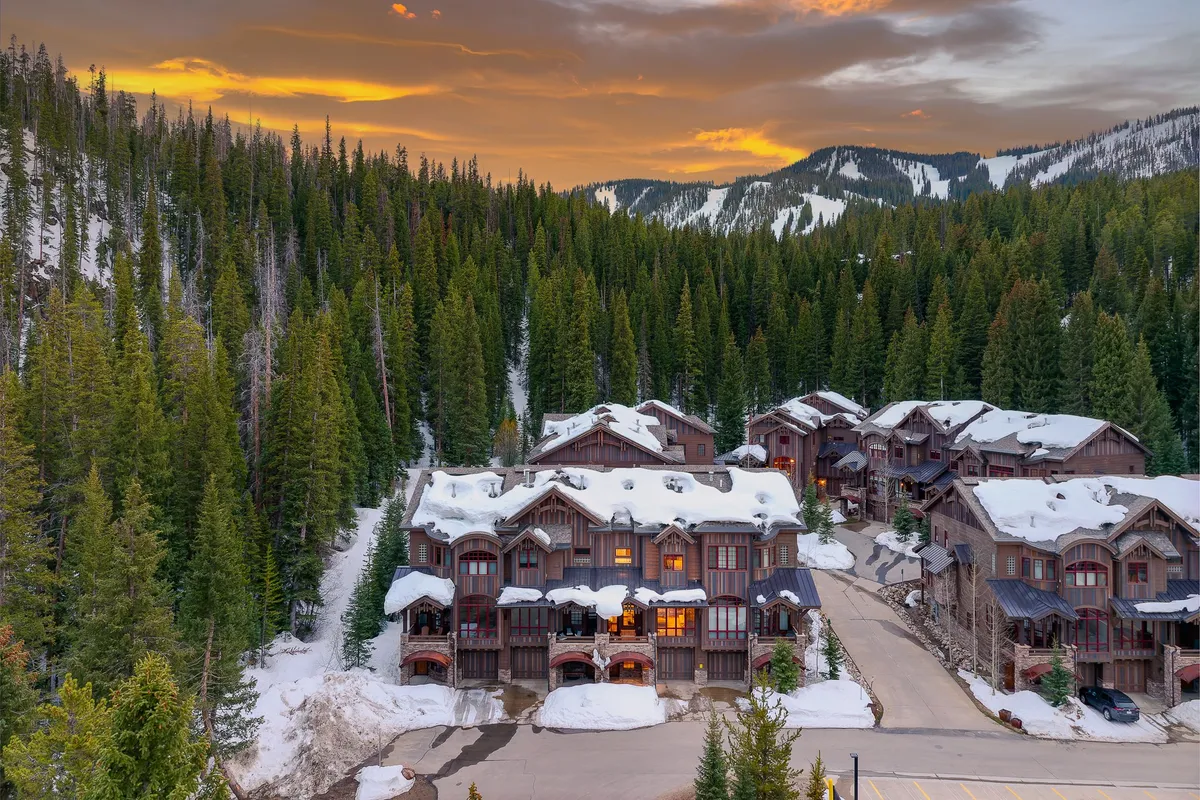 A picturesque view of snow-covered rooftops nestled among pine trees, with a mountain backdrop and glowing windows inviting guests inside after a day of exploration. The exterior setting is cozy yet adventurous, suggesting both comfort and easy access to the outdoors.