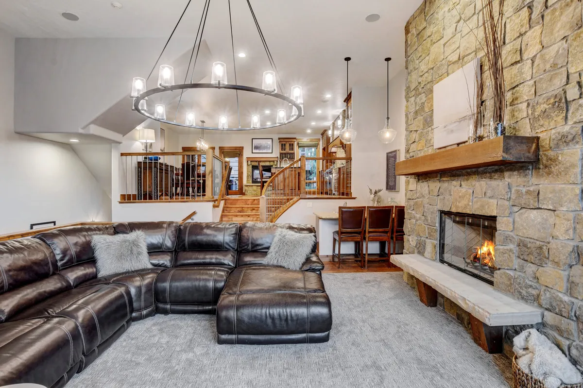 A spacious living room features a large leather sectional sofa positioned around a majestic stone fireplace, with a rustic chandelier overhead. The room is bathed in natural light from expansive windows that offer panoramic views of the mountains, and a wooden staircase adds architectural interest. The setup encourages gatherings, cozy movie nights, and lively conversations.