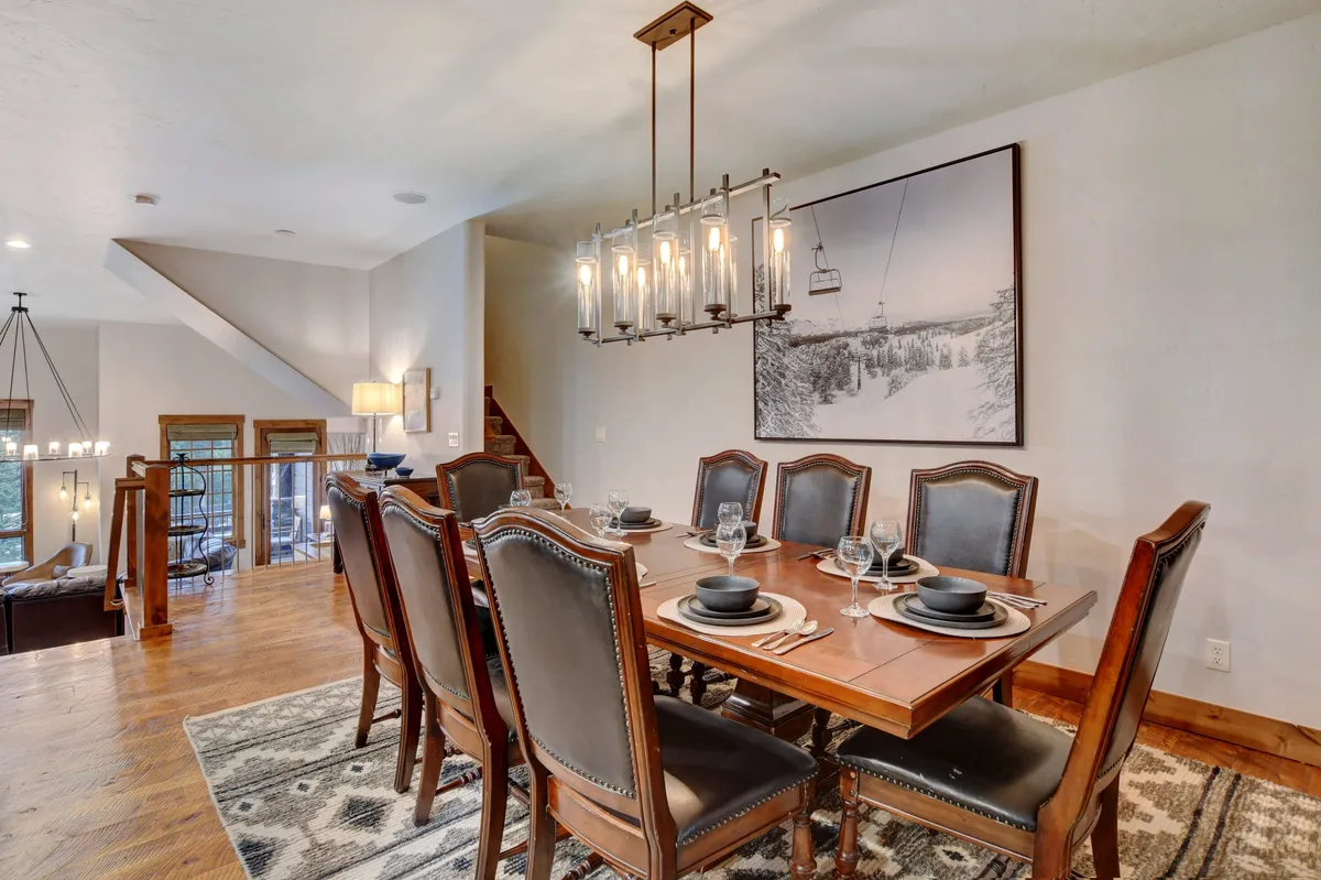 A warm dining area is centered around a massive wooden table under a striking chandelier. A mountain photograph adorns the wall, reinforcing the alpine setting, while the adjacent staircase hints at the open-concept layout. This space is ideal for festive family meals and heartfelt celebrations.