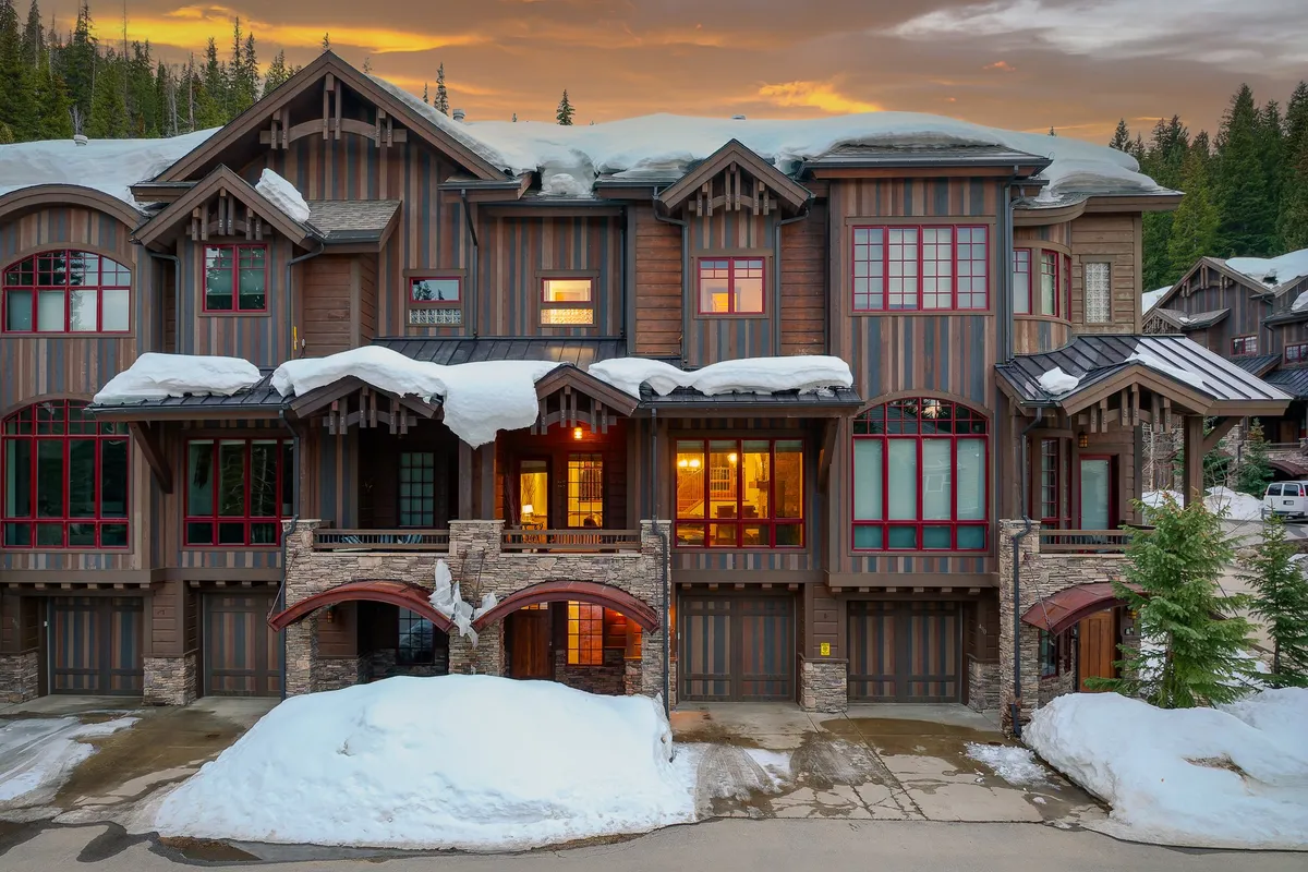 The image showcases the exterior of a luxurious single-family house in Winter Park, Colorado. Snow blankets the roof and grounds, while the home's wooden and stone facade glows with light from warmly lit windows. Majestic evergreen trees surround the property, creating a private mountain retreat. The inviting porch provides a perfect spot to watch sunsets over the snowy landscape, and the setting evokes cozy winter gatherings and outdoor fun.