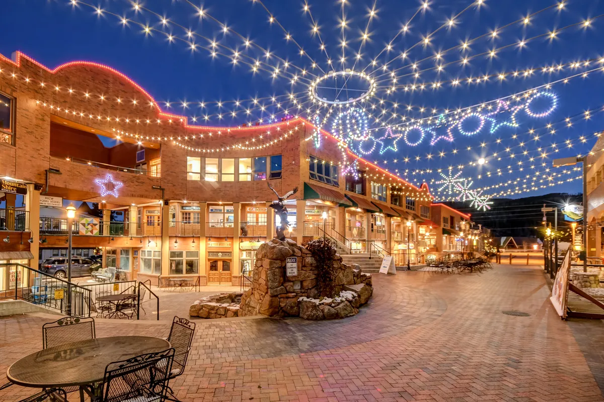 A cheerful exterior view of the resort village plaza, bustling with decorative lights, stone fountains, outdoor seating, and inviting storefronts. People are seen dining, shopping, and socializing, echoing the year-round activities and vibrant après-ski atmosphere described in the content.