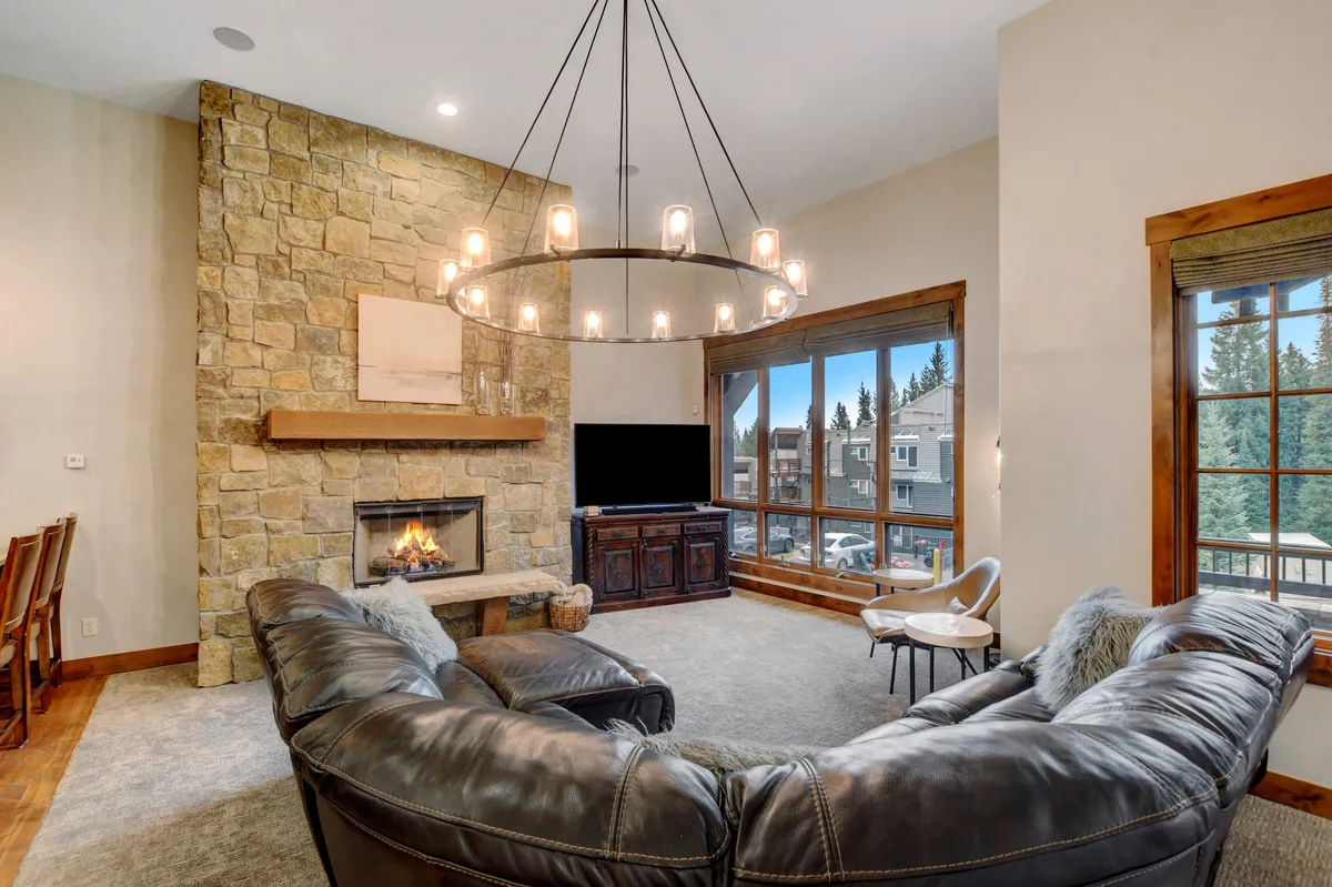 A spacious living area featuring a large sectional sofa, a stone fireplace, a chandelier, and expansive windows with scenic mountain views. The room radiates warmth and comfort, perfect for cozy family gatherings or quiet evenings by the fire.