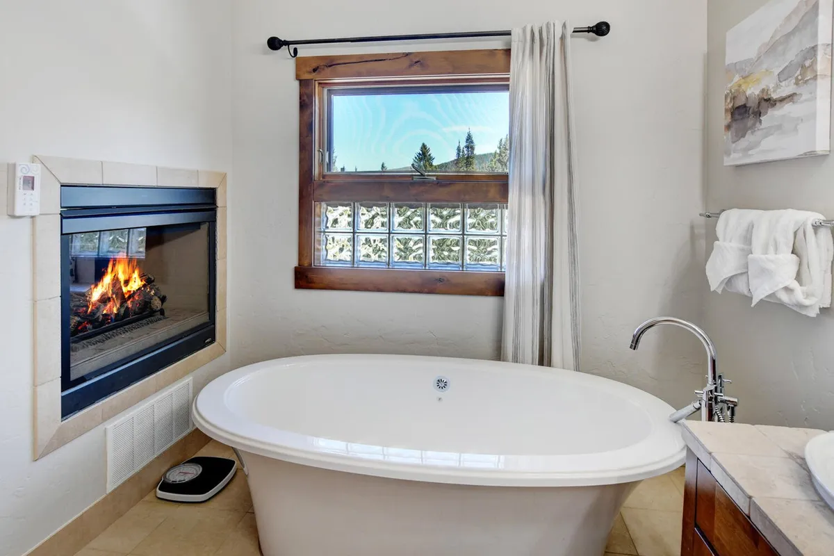A luxurious bathroom featuring a freestanding bathtub adjacent to a fireplace, with a window offering an outdoor view. The space is serene and intimate, perfect for unwinding or sharing a quiet moment.